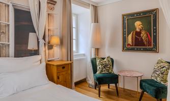 A cozy bedroom with a large bed, a nightstand, and two green chairs. A striking painting hangs on the wall.