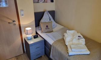 A cozy single room with a bed, a nightstand, and a chair. The walls are painted in soft colors, and there are fresh towels on the bed.