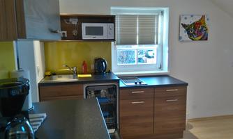 A modern kitchen with wooden cabinets and a yellow wall. There is a microwave, a kettle, and a window with a view outside.