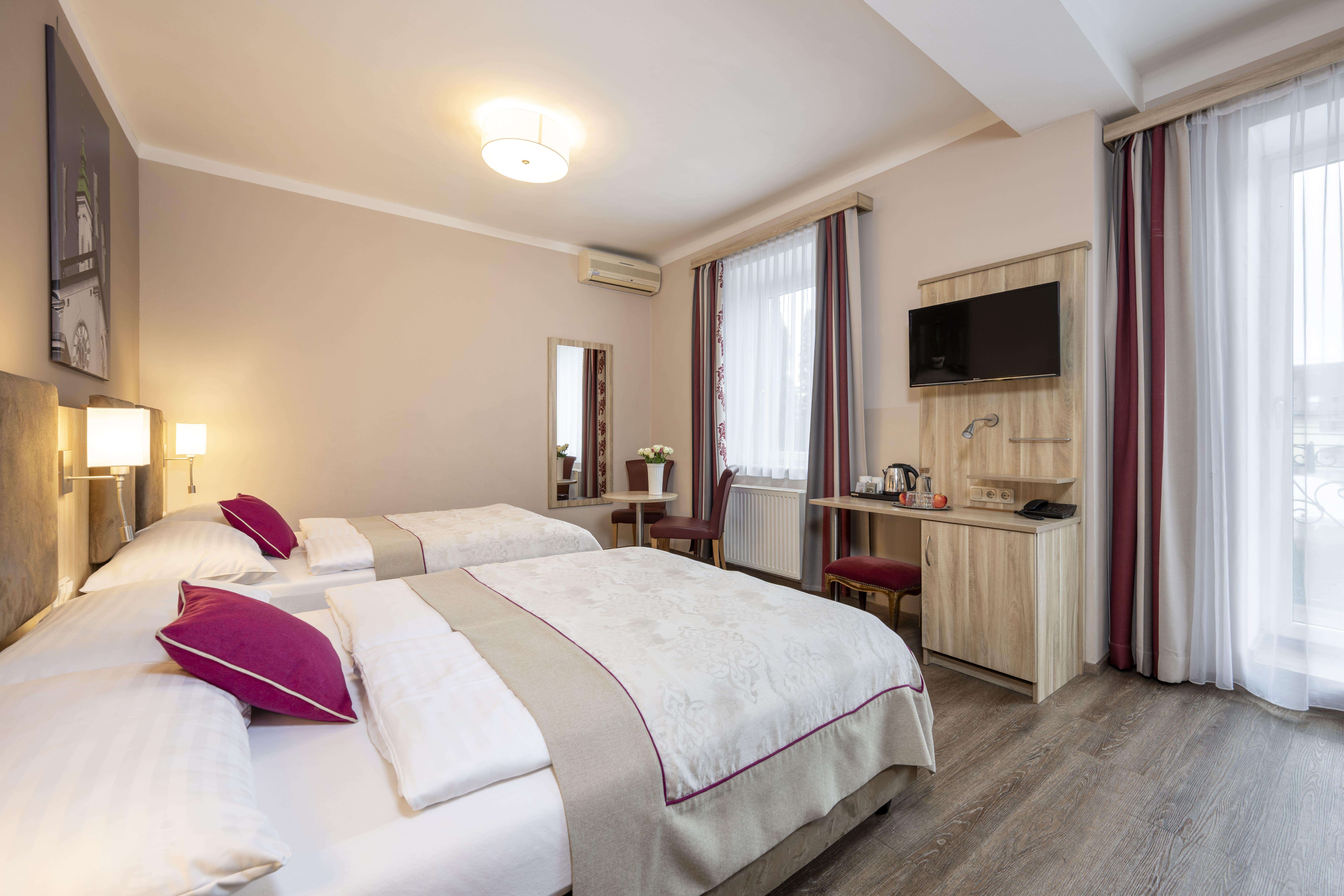 A bright hotel room with two single beds and modern decor. It features a table, a television, and large windows that let in plenty of light.