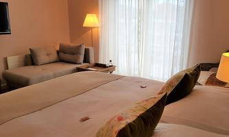 A cozy hotel room with two beds and a bright living area. Large windows let in plenty of light and create an inviting atmosphere.