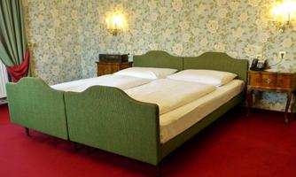A cozy bedroom with a green double bed and a red carpet. The walls are decorated with floral designs and there is a nightstand in the room.