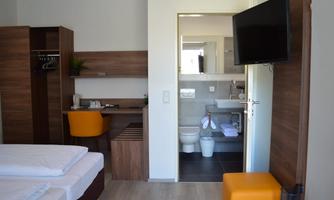 A modern hotel room with two single beds, a desk, and a flat-screen TV. In the background, a bathroom with a toilet and sink is visible.