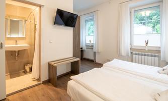 A modern room with two beds and a flat-screen TV. In the background, a bathroom with a shower is visible.