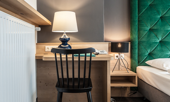 A cozy room with a wooden desk and a black chair. An elegant bedside table with two lamps and a large green headboard.