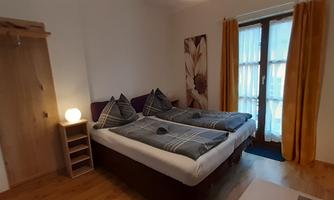 A cozy bedroom with two single beds and a warm color scheme. Large windows with curtains let in plenty of daylight.