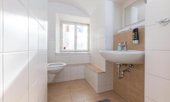 A modern bathroom with white tiles and a large window. It includes a sink, a toilet, and a cozy seating area.