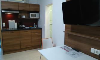 A modern kitchen unit with a wood look and appliances such as a microwave and coffee machine. A small table with a chair and a television are also visible in the room.
