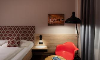 A modern hotel room with a red chair and a small table. The walls are in soft colors, and the bed has a decorative headboard.