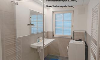 A modern bathroom with a shower, sink, and window. There is a washing machine and towel holders on the wall.