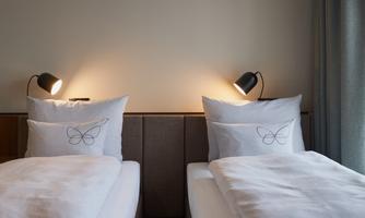 A modern bedroom with two single beds and elegant bedside lamps. The pillows have a simple butterfly design.