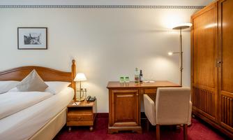 A cozy hotel room with a double bed and a wooden desk. There is a lamp, a chair, and a small dresser.