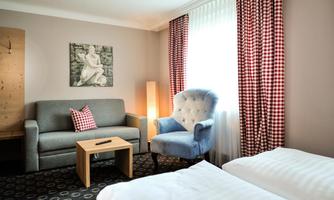 A cozy hotel room with two beds, a gray sofa, and a blue armchair. Bright curtains and a piece of art on the wall create an inviting atmosphere.