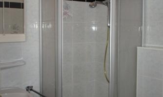 A small shower with white tiles and a glass partition. In the room, there is a mirror and a hand shower visible.