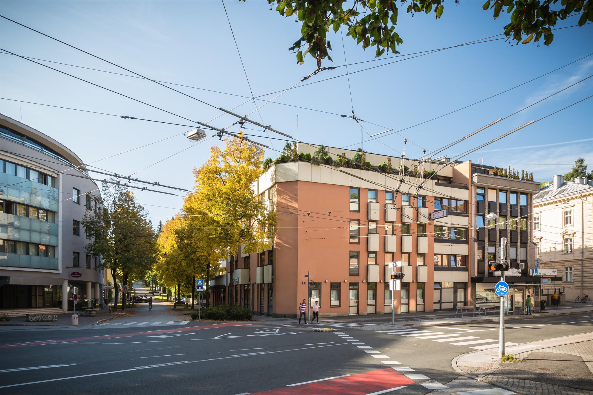 A modern building at a street corner, surrounded by trees and a clear sky. Tram lines stretch across the street.