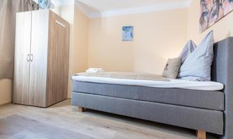 A simple room with a single bed and gray bedding. Next to the bed stands a wooden wardrobe and on the wall hangs a small picture.