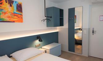 A modern room with a double bed, a nightstand, and a mirror. A colorful painting hangs on the wall, and there is a slim wardrobe.