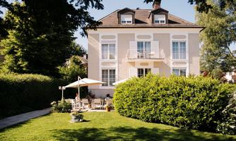 An elegant house surrounded by a well-maintained garden. On the porch, there are tables and chairs for cozy hours outdoors.