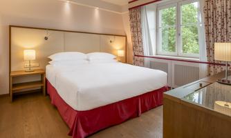 A cozy hotel room with a large bed and wooden floor. The room has two windows that let in plenty of daylight.