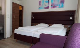 A modern room with two single beds and a purple sofa. Colorful paintings hang on the wall, and there is a wardrobe.
