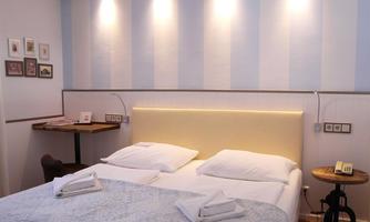 A bright bedroom with a large double bed and elegant lighting. Next to the bed is a small table with a chair.