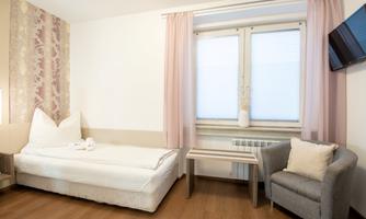 A simple room with a bed, a gray armchair, and a small table. Large windows with light curtains let in a lot of light.