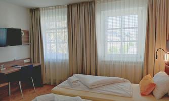 A modern hotel room with two beds and a desk. Large windows let in plenty of daylight.