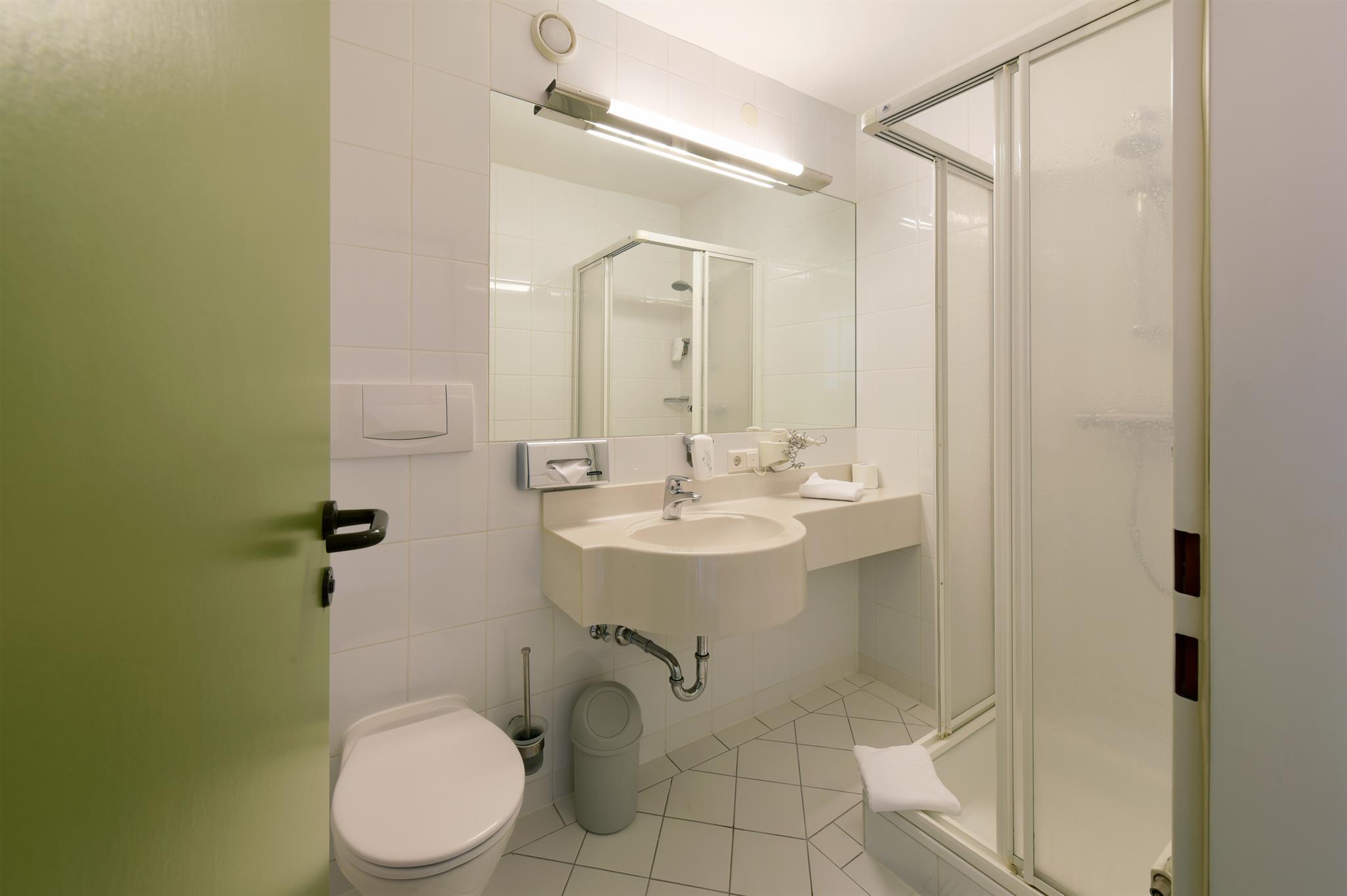 A modern bathroom with a shower, sink, and mirror. The walls are tiled white, and the door is green.