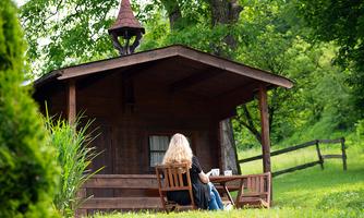 A cozy little house in nature, surrounded by green trees. A person is sitting relaxed on a bench, enjoying the view.