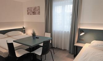 A modern room with two beds and a small table. Large windows let in plenty of daylight.