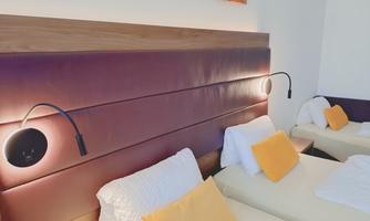 A modern room with two single beds and decorative white pillows. The wall behind the bed is painted in dark purple and features integrated bedside lamps.