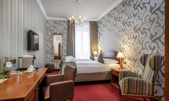 A cozy hotel room with two beds and elegant wallpaper. There is a television, a table with chairs, and plenty of daylight coming through the window.