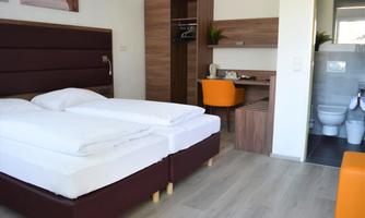 A modern hotel room with two single beds, a desk, and a bathroom in the background. The decor is simple and functional, with warm tones.