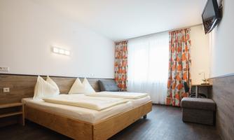 A modern bedroom with a double bed, wooden furniture, and colorful curtains. There is a small table, a chair, and a TV on the wall.
