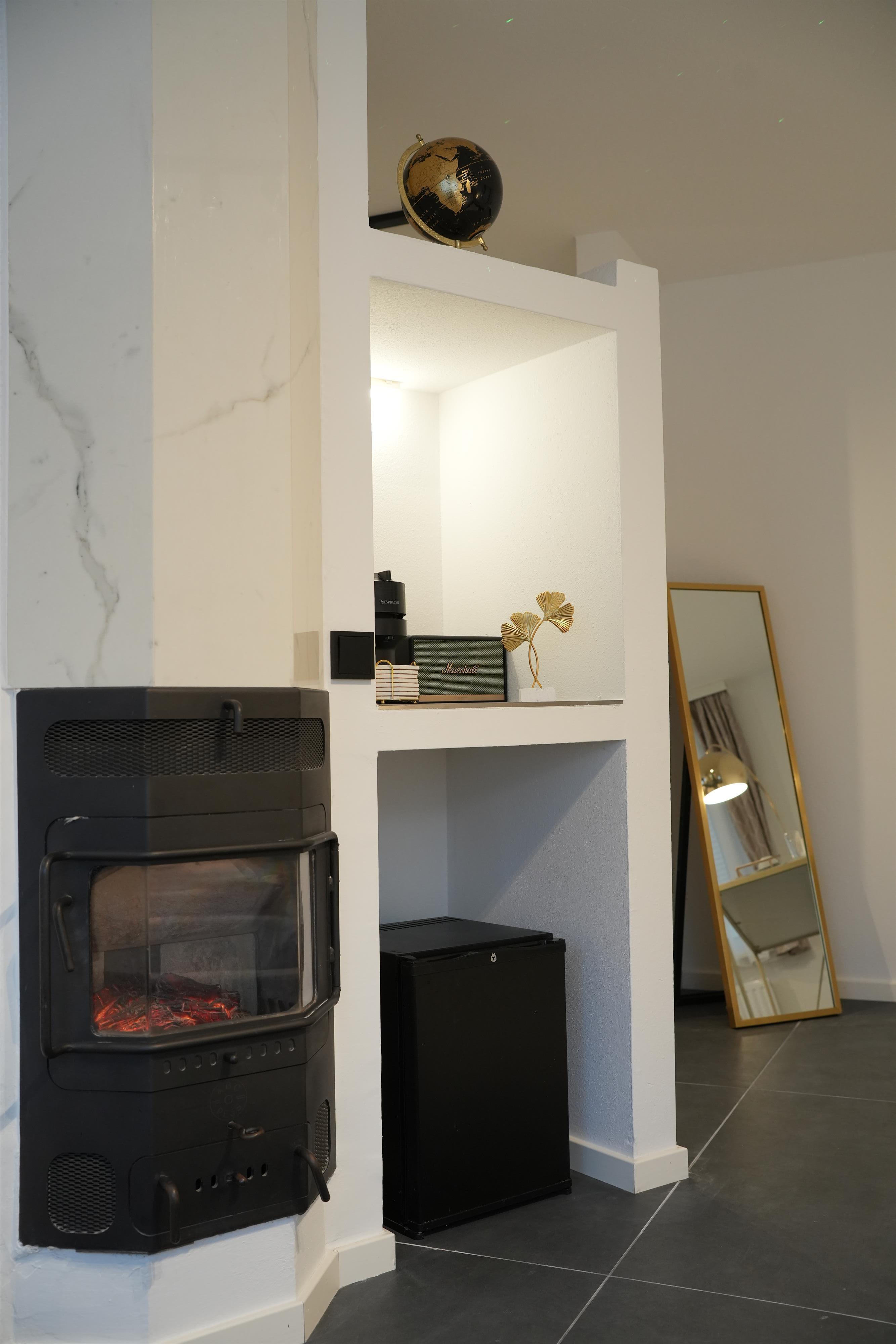 A cozy living space with a modern stove, a small refrigerator, and a golden table lamp. In the corner stands a large mirror that brightens the room even more.