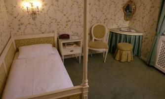 An elegant bedroom with a single bed, a dresser, and a vanity table. The walls are decorated with floral patterns, and there is a stylish chair.