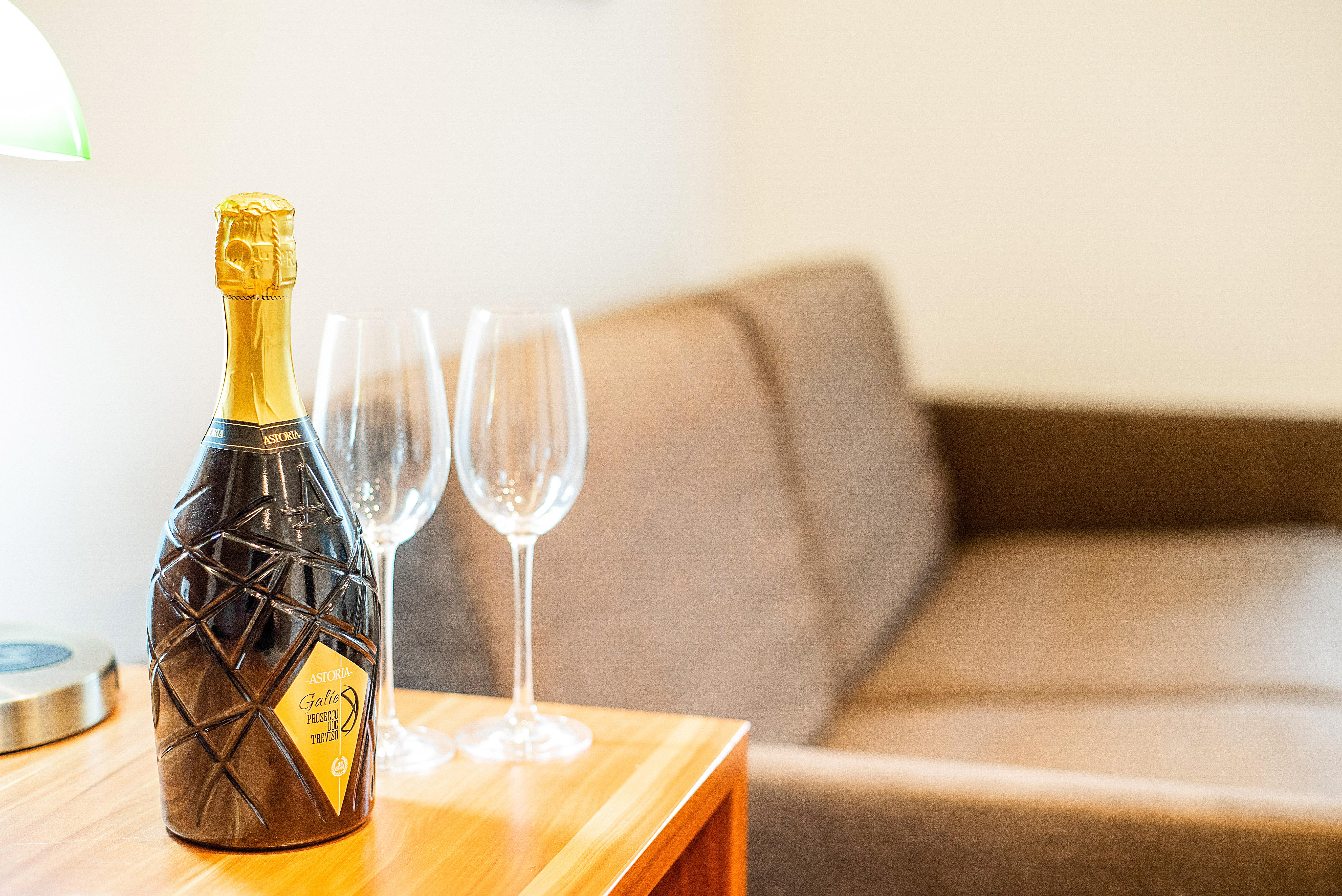 A bottle of sparkling wine is on a wooden table next to two glasses. In the background, a cozy sofa can be seen.