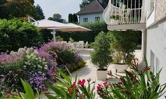 A beautiful garden with colorful flowers and green plants. In the background, a cozy terrace with a sun umbrella is visible.