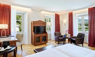 An elegant hotel room with two single beds, a cozy seating area, and a television. Large windows let in plenty of light and offer a view of the surrounding nature.