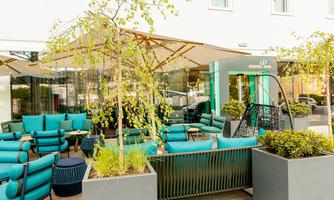 A modern terrace with comfortable turquoise furniture and plants. In the background, the entrance area of a hotel can be seen.