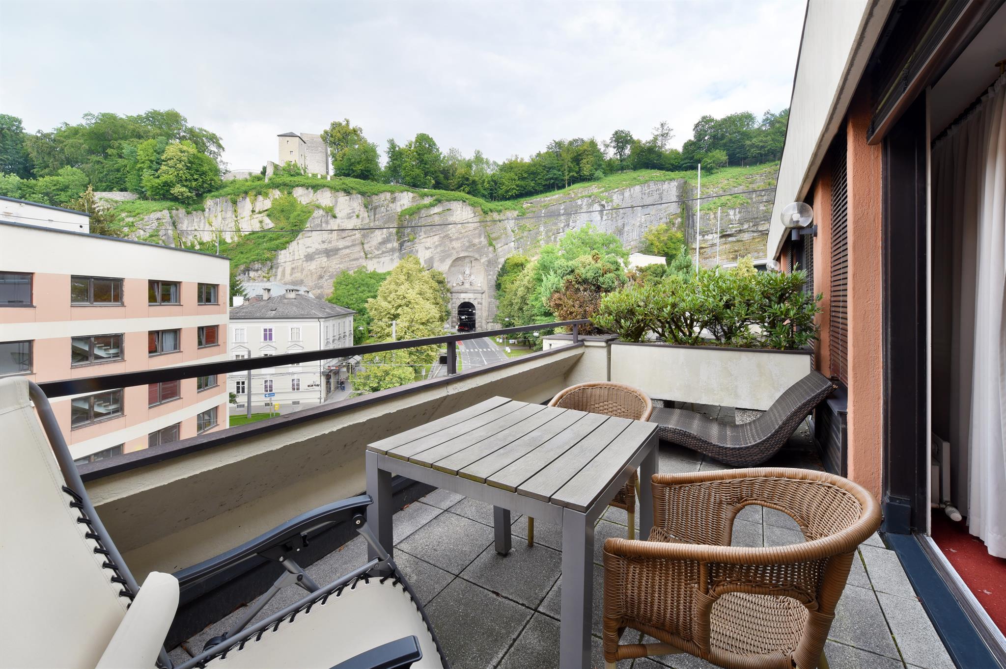 A cozy balcony with a table and chairs. In the background, green hills and an older building complex can be seen.