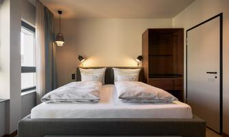 A modern bedroom with a comfortable double bed and soft lighting. The walls are light, and a wardrobe is visible in the background.