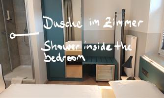 A modern bedroom with a double bed and a shower in the room. The walls are painted in light colors, and there is a small table and a chair.