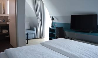 A modern hotel room with two beds and a television. There is a cozy armchair and curtains that gently filter the light.