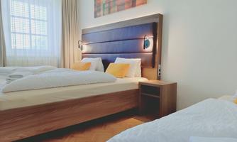 A modern bedroom with two beds and a cozy wooden frame. The room has large windows with curtains and indirect lighting.