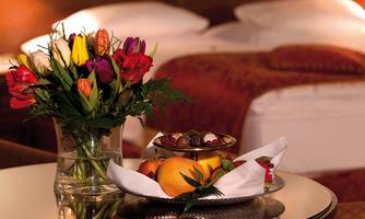 A table with fresh flowers and a bowl full of fruit and chocolates. In the background, two beds can be seen in an elegant room.