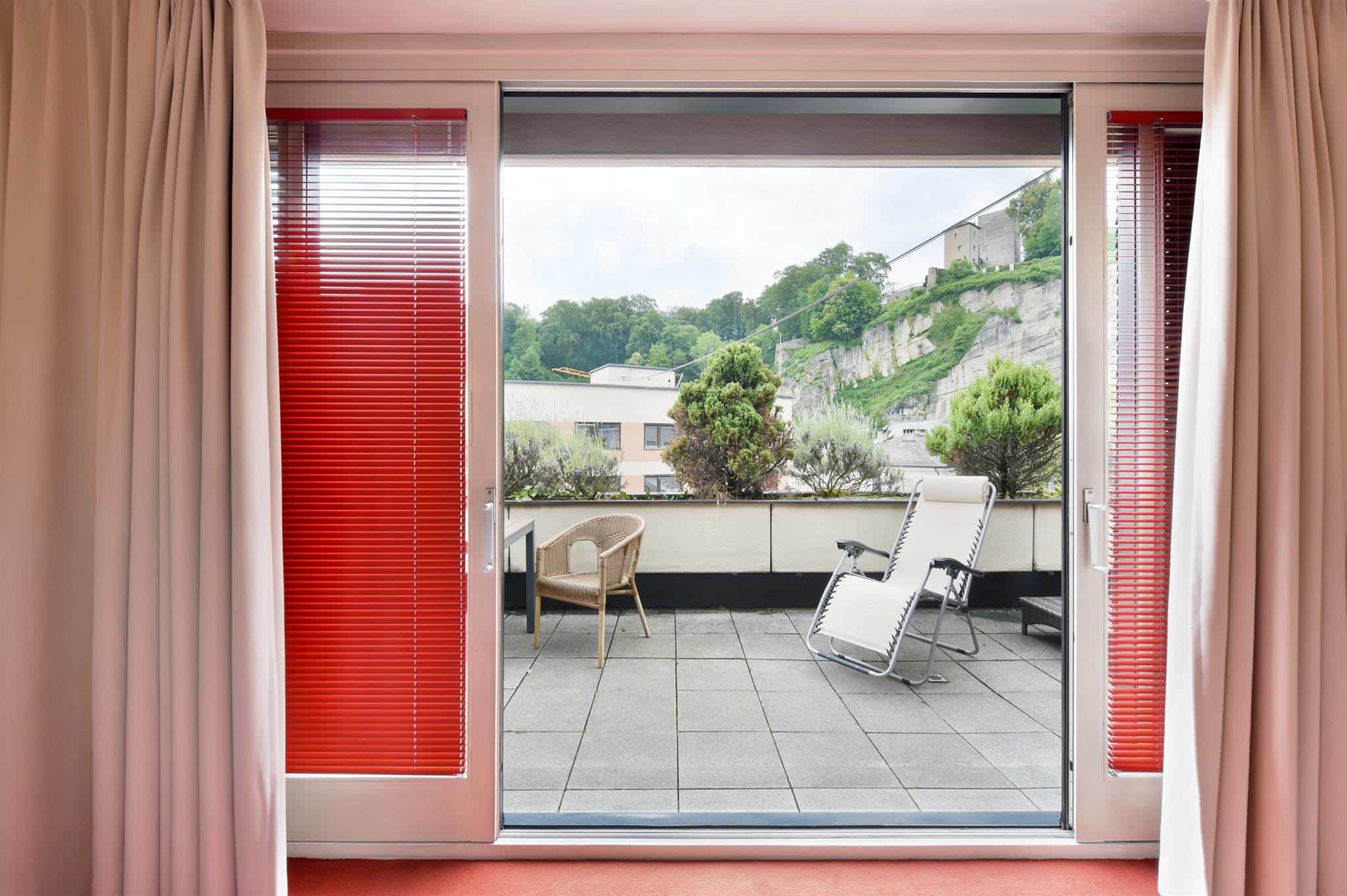 A modern room with a view of a terrace. The windows have red blinds and there is a chair and a lounge on the balcony.