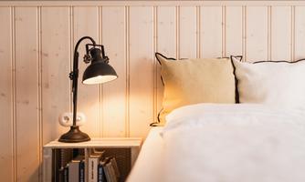A cozy bedroom with a bed, a lamp, and decorative pillows. The wooden wall paneling creates a warm atmosphere.