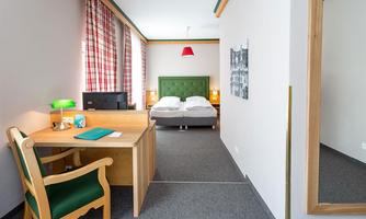A cozy hotel room with a double bed and a desk. The walls are painted in soft colors, and the light creates an inviting atmosphere.