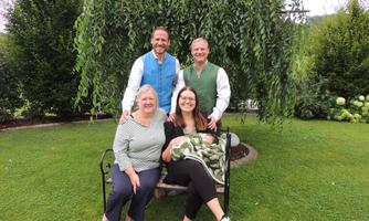 A group of four people is posing outdoors on a bench. In the background, there are large trees and green grass.